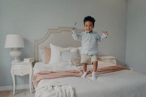Joyful small afro american kid jumping on bed at home and smiling photo