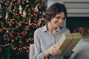 Happy young Italian woman sits on cozy sofa next festive decorated Xmas tree with Christmas present photo