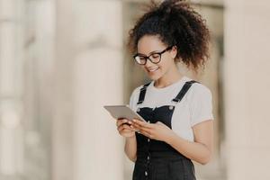 Pretty curly haired girl looks at digital tablet screen with smile, reads some text or electronic book, wears spectacles for vision correction, white t shirt and dungarees, enjoys high speed internet photo