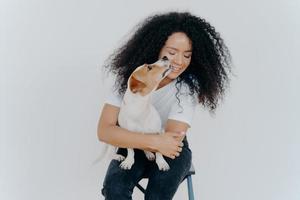 Cute happy woman with curly hair gets kiss from jack russell terrier feels love to favourite pet takes pleasure in company of dog sits on chair against white background. Love between owner and animal.