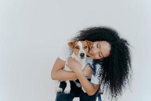 Image of playful pretty woman with Afro hairstyle, smiles gladfully, holds domestic animal, carries dog to veterian, isolated over white background. Glad lady petts jack russell terrier at home. photo