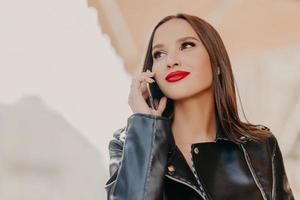 Headshot of pleasant looking dreamy woman focused aside, has telephone conversation, wears leather coat, notices someone into distance, poses over blurred background. Good roaming connection photo