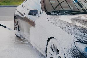 Black dirty car in white soap foam at car wash service station photo