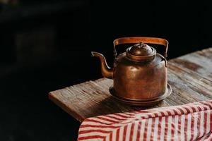 Horizontal shot of antique kettle on vintage wooden table. Old crockery. Metal copper teapot against dark background red towel near. Selective focus photo