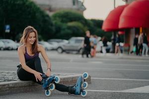 estilo de vida deportivo y afición. una mujer europea de pelo oscuro complacida se pone patines en línea haciendo poses de patinaje contra el fondo borroso de la ciudad se mantiene en forma pasa el tiempo libre activamente. tiro al aire libre
