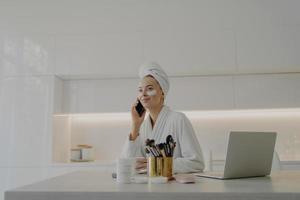 Beautiful woman in bathrobe with towel on head talking on mobile phone while doing skin care procedures at home photo