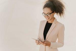 Happy business lady stands with touchpad, reads news online, wears spectacles and elegant clothes, has curly hair combed in pony tail, stands over white wall with blank space, looks for information photo