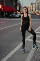 una foto completa de una joven activa que está en buena forma física vestida con ropa deportiva negra disfruta patinando durante las buenas poses del día de verano en la carretera contra el fondo de la ciudad. recreación.