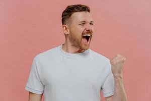 Emotional successful man shouts from joy clenches fist with triumph makes pump gesture cellebrates triumph dressed in casual white t shirt isolated over pink background. Body language concept photo