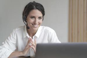 Smiling italian businesswoman in wireless headphones sitting at desk while works on laptop at home photo