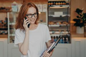 Happy ginger shop owner has telephone conversation holds mobile phone near ear photo