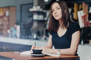 Concentrated female journalist thinks about new article, writes down information into notepad, drinks hot aromatic beverage in coffee shop, modern smart phone on table near. Working process. photo