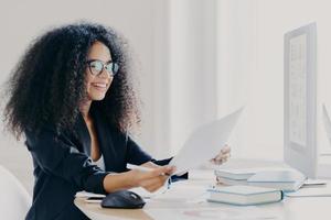 Glad Afro American woman with curly hairstyle, makes financial report, holds paper documents, has happy smile wears spectacles for vision correction dressed in elegant clothes computer screen in front photo
