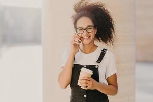 una adolescente rizada de aspecto agradable usa anteojos transparentes, habla por teléfono móvil, toma un descanso para tomar café, se viste con una camiseta blanca y un mono, disfruta del tiempo libre y la conversación. gente y tecnología foto