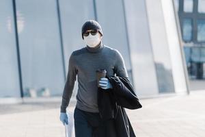 Image of serious man walks to work, wears hat, sunglasses, drinks coffee to go, holds newspaper, avoids public place during quarantine time, poses outdoor. Covid-19, virus concept. Influenza photo