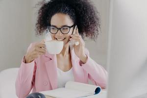 Cropped image of happy young African American woman makes phone call, drinks aromatic latte or espresso, poses in office interior, has pleasant smile wears optical glasses and rosy formal jacket photo