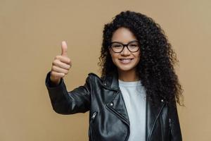 I totally agree with you. Pleased curly haired woman shows thumb up, rates excellent product, recommends promo offer, wears glasses and leather jacket, isolated on beige background, approves best deal photo