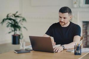 Serious bearded man writes informative notes while watching webinar on laptop computer works remotely dressed in casual t shirt poses at workplace against home office interior. Modern workspace photo