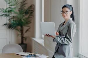 Woman administrative stands near desktop, holds modern laptop computer, uses wireless connection to internet, dressed in formal grey outfit, searches information on web page, works distantly photo