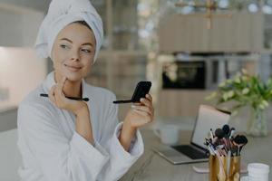 Tender young woman has morning routine applies daily makeup holds cosmetic brush and mirror touches gently face wears bathrobe and towel on head poses against home interior enjoys skin care. photo