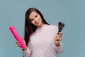 Pleased young female model makes hairstyle and makeup, holds bottle of hairspray and cosmetic brushes, tilts head, dressed in elegant clothes, poses over blue background. Beauty and style concept photo