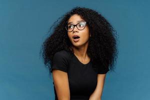 Impressed dark skinned lady with frizzy hair, looks surprisingly aside, keeps jaw dropped, notices something astonishing, wears optical glasses and casual black t shirt, isolated over blue background photo