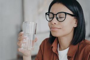 Happy woman wearing glasses holds glass of pure water, closed eyes. Healthy lifestyle, wellness photo