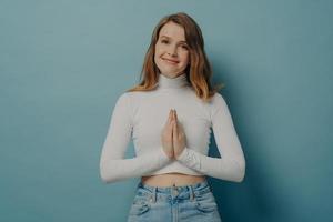 Young calm female keeping hands folded in namaste gesture while standing in front of blue wall photo