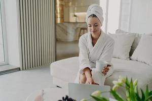 Young woman in white bathrobe working remotely on laptop while sitting on sofa in modern living room photo