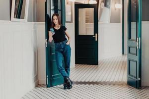 Full length shot of serious fashionable woman concentrated into cell phone, wears black t shirt, jeans and boots, reads information in internet, stands indoor near doors connected to wireless internet photo