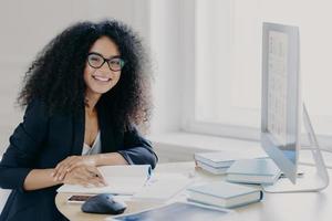 un oficinista alegre y rizado ve un seminario web sobre un tema de negocios, escribe notas con un bolígrafo, sonríe positivamente, usa anteojos y ropa negra prepara un informe con anteojos transparentes. espacio de trabajo conjunto foto