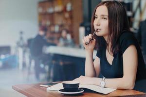 toma interior de una joven estudiante que escribe notas para el informe, trabaja en una cafetería, bebe capuchino, planifica el proceso de organización, tiene una expresión reflexiva, crea un ensayo o se prepara para el examen foto