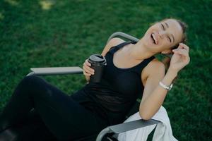 Overjoyed relaxed woman smiles broadly enjoys drinking coffee outdoor dressed in t shirt and trousers poses in comfortable chair against green grass. View from above. People leisure lifestyle photo