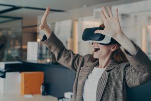 Impressed red-haired business woman dressed wearing portable VR goggles exploring virtual world photo