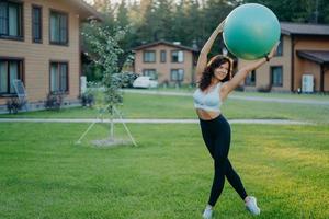 Sporty slim woman wears cropped top and leggings, has aerobics exercises with fitness ball, poses outdoor on green lawn, private houses in background. Healthy fit female model raises fitball over head photo
