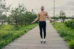 Sporty muscular man uses skipping rope for cardio training outdoor, has perfect body, breathes fresh air and demonstrates endurance, works out self development. Athlete guy with sport equipment photo
