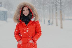 Outdoor shot of attractive woman dressed in winter clothes, keeps both hands in pockets, looks with satisfied expression directly at camera, walks in open air during snowfall. Snowy weather. photo