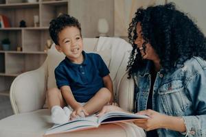 encantadora madre afroamericana leyendo un libro a su pequeño hijo y riendo foto