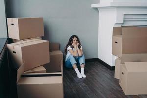 Frustrated woman is looking at packed boxes sadly and sitting on floor. Girl is upset with moving. photo