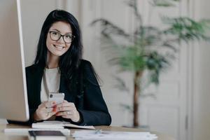 Happy female office worker in stylish black jacket browses smartphone with cheerful expression, sends text messages, works on important project, poses in coworking space. Modern technologies photo