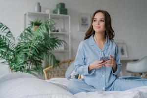 Shot of pensive female model uses smart phone and earphones for watching movie, connected to wireless internet, dressed in striped casual nightwear, poses in lotus pose on bed, has good rest