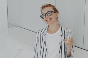 Young beautiful pleased red-haired woman having healthy habits, drinking mineral water in morning photo