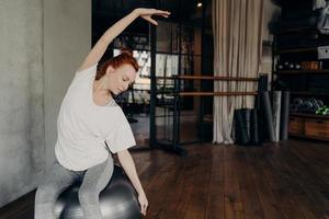 mujer pelirroja deportiva sentada en un gran fitball y estirando el cuerpo antes de hacer ejercicio en el gimnasio foto