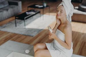 vista desde arriba de una hermosa mujer relajada y satisfecha cierra los ojos y disfruta de la suavidad de la piel, tiene tratamientos de belleza después de la ducha, sostiene una botella de loción, posa en casa, siente comodidad y placer foto