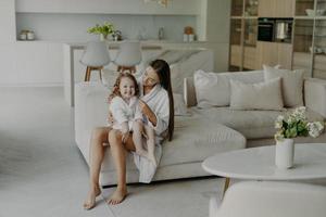 Happy adorable little hands poses on mothers legs dressed in soft white bath towel feels very glad. Mom and daughter wear domestic clothes sit on comfortable sofa in living room. Home interior photo