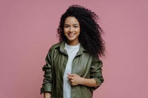 foto de media longitud de una mujer afroamericana positiva vestida con pantalones de cuero, sonríe agradablemente, disfruta de un buen día, pasa el fin de semana con amigos, tiene el pelo rizado y tupido, aislada en un fondo rosado.