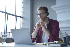 Pensive German businessman in headphones sits at desk while remotely working from home photo