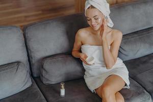 Top view of pleased healthy woman sits on comfortable couch indoor, applies face cream and uses body lotion after taking bath, has natural makeup and healthy skin. Hygiene and beauty concept photo