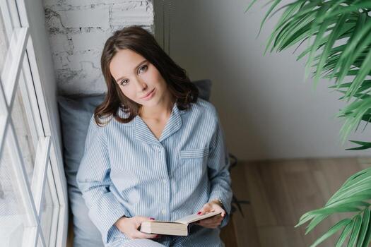 Top view of relaxed pleasant looking woman enjoys reading book, dressed in casual domestic suit, sits on window sill for good light and vision, enjoys loneliness and spare time. Hobby concept photo