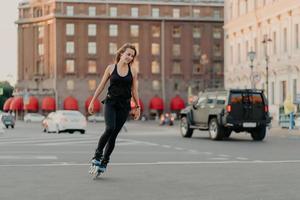 Active girl rollerblading on grey asphalt poses on rollers dressed in active wear drives fast leads healty lifestyle enjoys favorite hobby. Sport recreation roller skating summer time concept photo